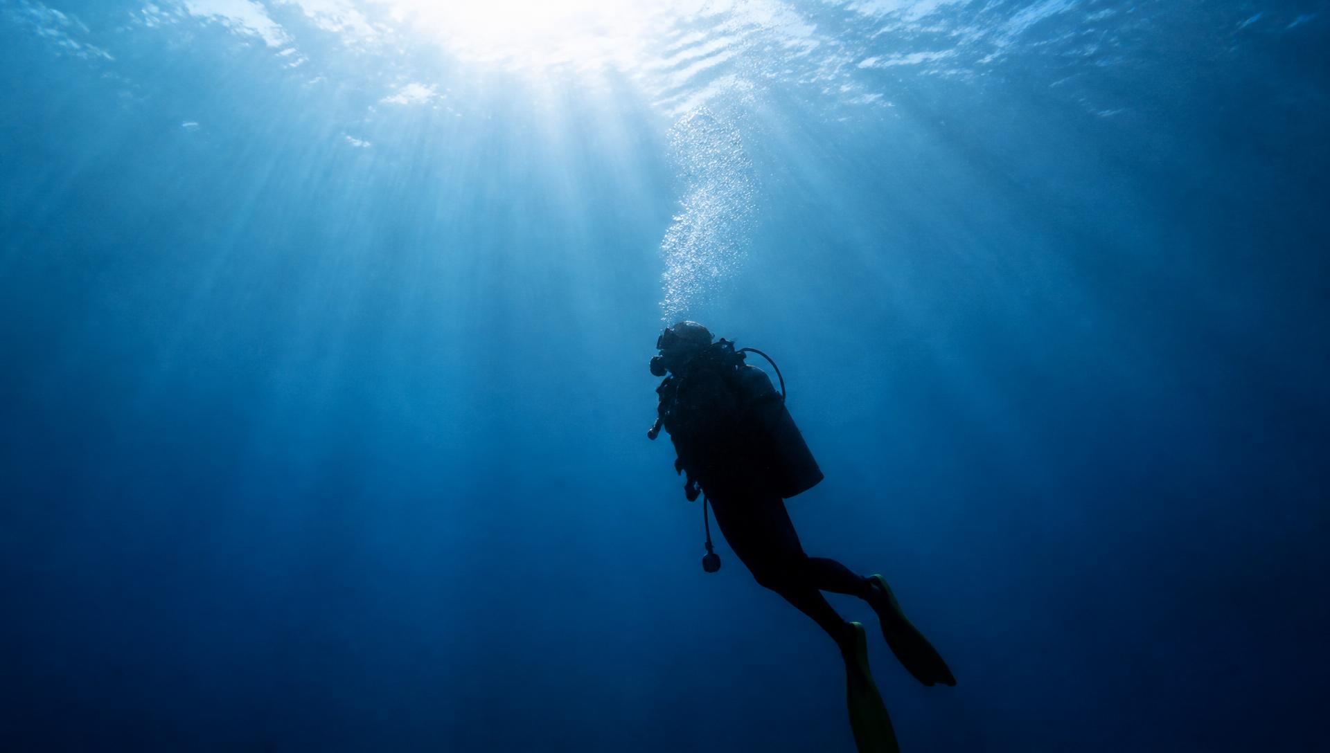 Lone scuba diver silhouetted in deep blue water with sunlight from the surface above
